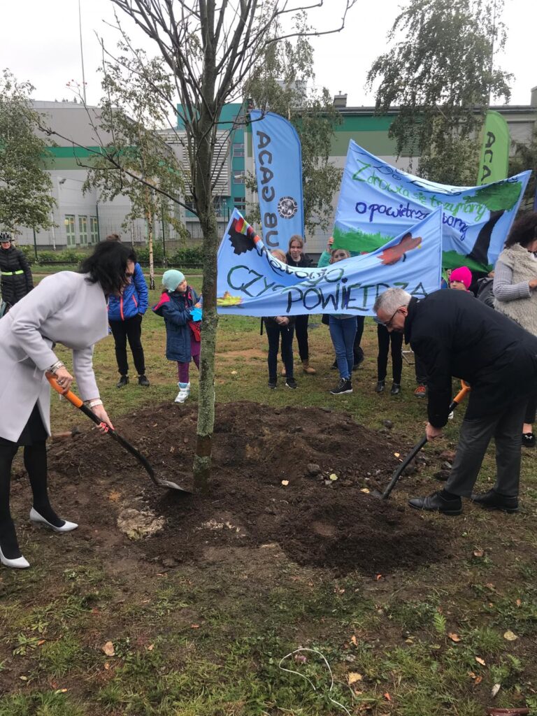 Na pierwszym planie kanclerz Agnieszka Skołucka oraz Dyrektor Jan Siechowski zasadzający nowe drzewo przed budynkiem CINiB-y. Na drugim planie dzieci z transparentami dotyczącymi dbania o czyste powietrze.