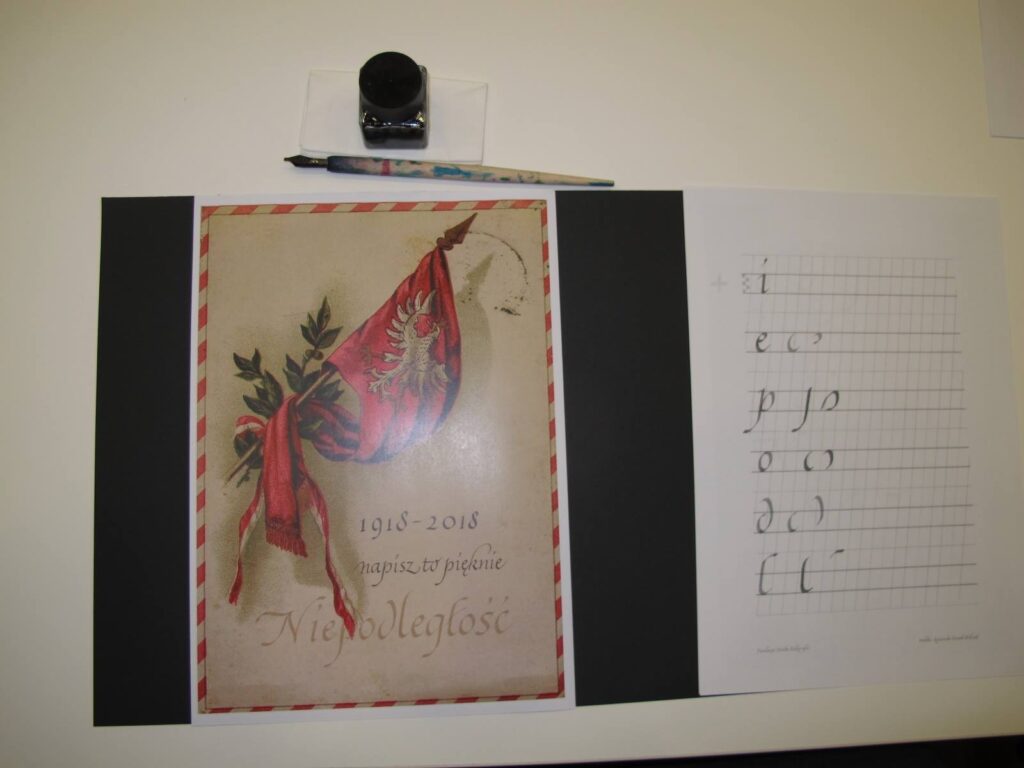 Na zdjęciu zeszyt do nauki kaligrafii oraz szklany pojemnik z atramentem oraz pióro.