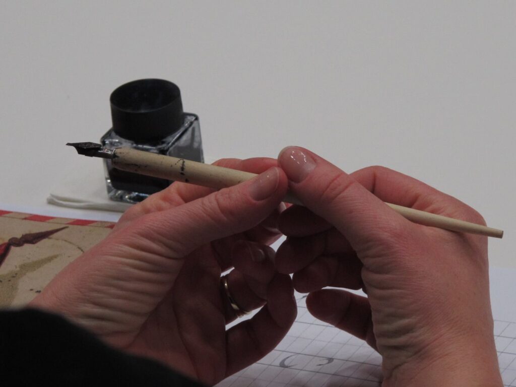 Na pierwszym planie zbliżenie na dłonie, które trzymają pióro ubrudzone z atramentu, a na drugim planie szklany pojemnik z atramentem.
